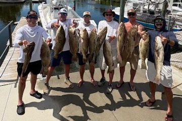 Six people hold large fish on a dock with boats in the background.