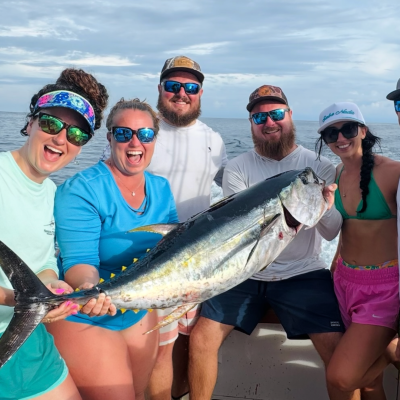 Six people on a boat holding a large fish with fishing rods, smiling against a backdrop of ocean and cloudy sky.
