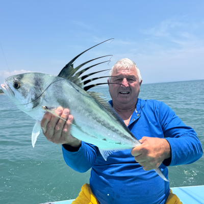 Person holding a roosterfish on a boat with fishing rods and ocean background.